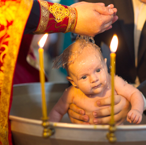 A priest uses his hands to pour Holy Water on the head of a baby in a round baptismal fount.